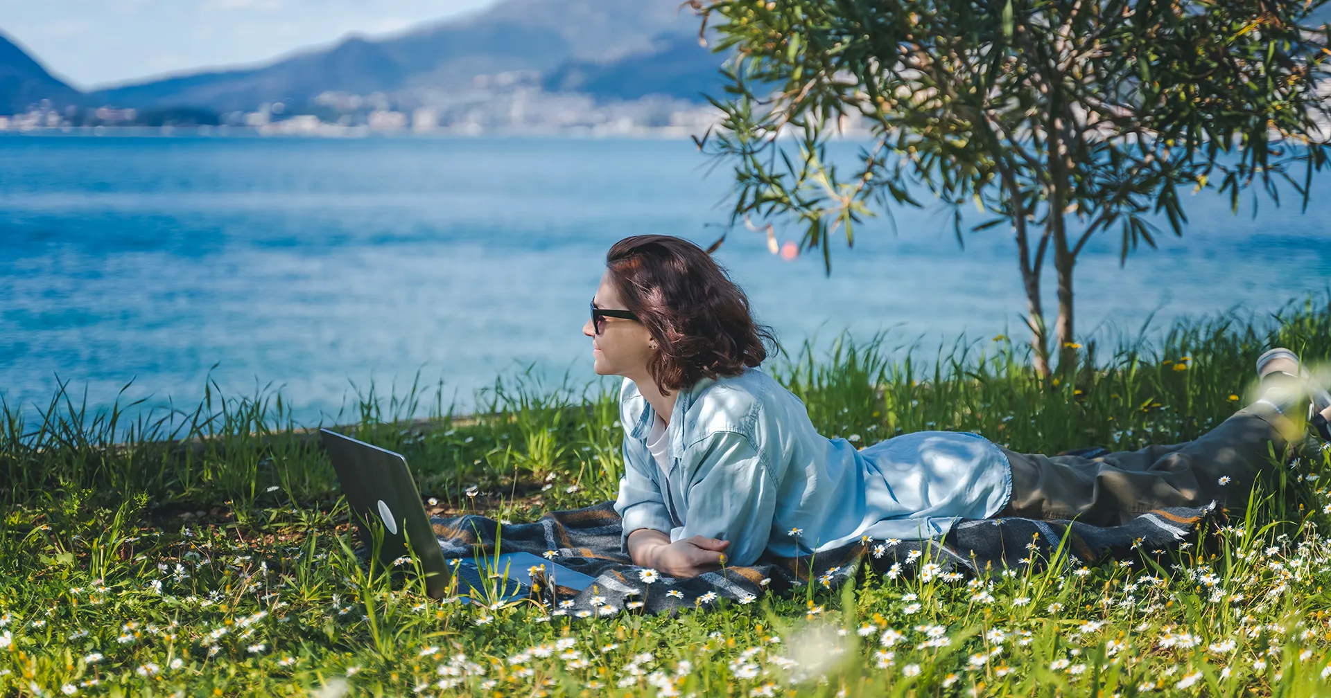 Vrouw is op vakantie en ligt op haar buik in het gras achter haar laptop aan het water